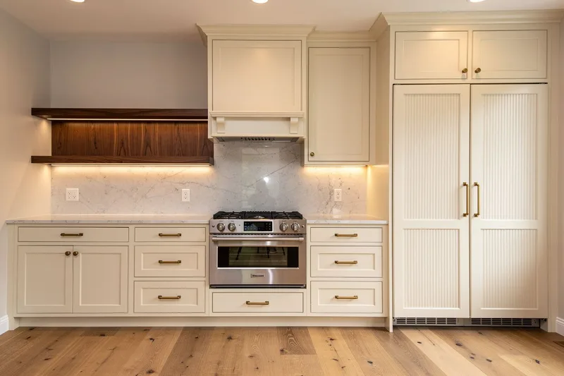 Cream shaker kitchen cabinets with brass cup pulls, a paneled range hood, tall pantry cabinets and honed marble counters